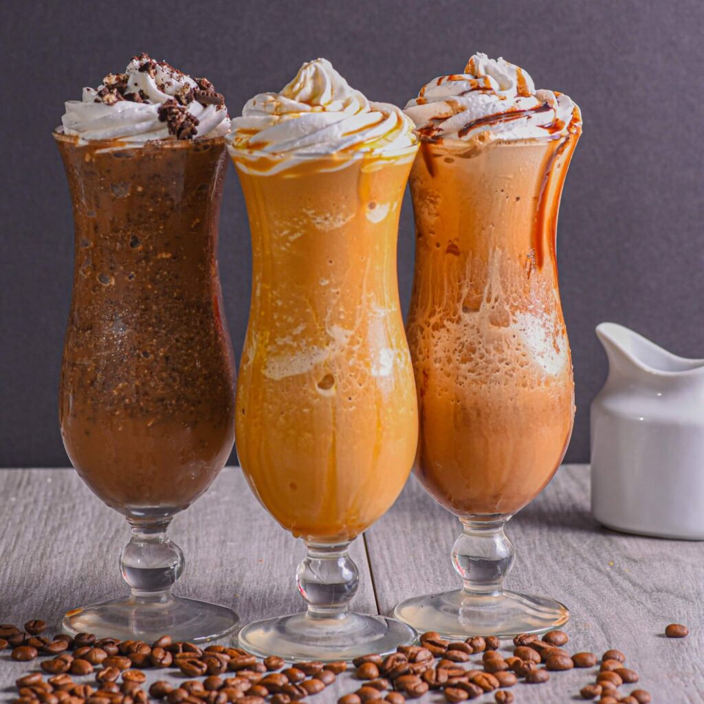 On a table, three coffee smoothies with whipped cream and coffee beans below.